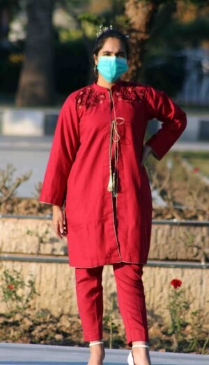 A woman standing in a garden wearing a elegant 3-piece crimson Dori Affairn Pakistani-style suit with shoulder embroidery, gold piping, and a decorative tassel dori closure.